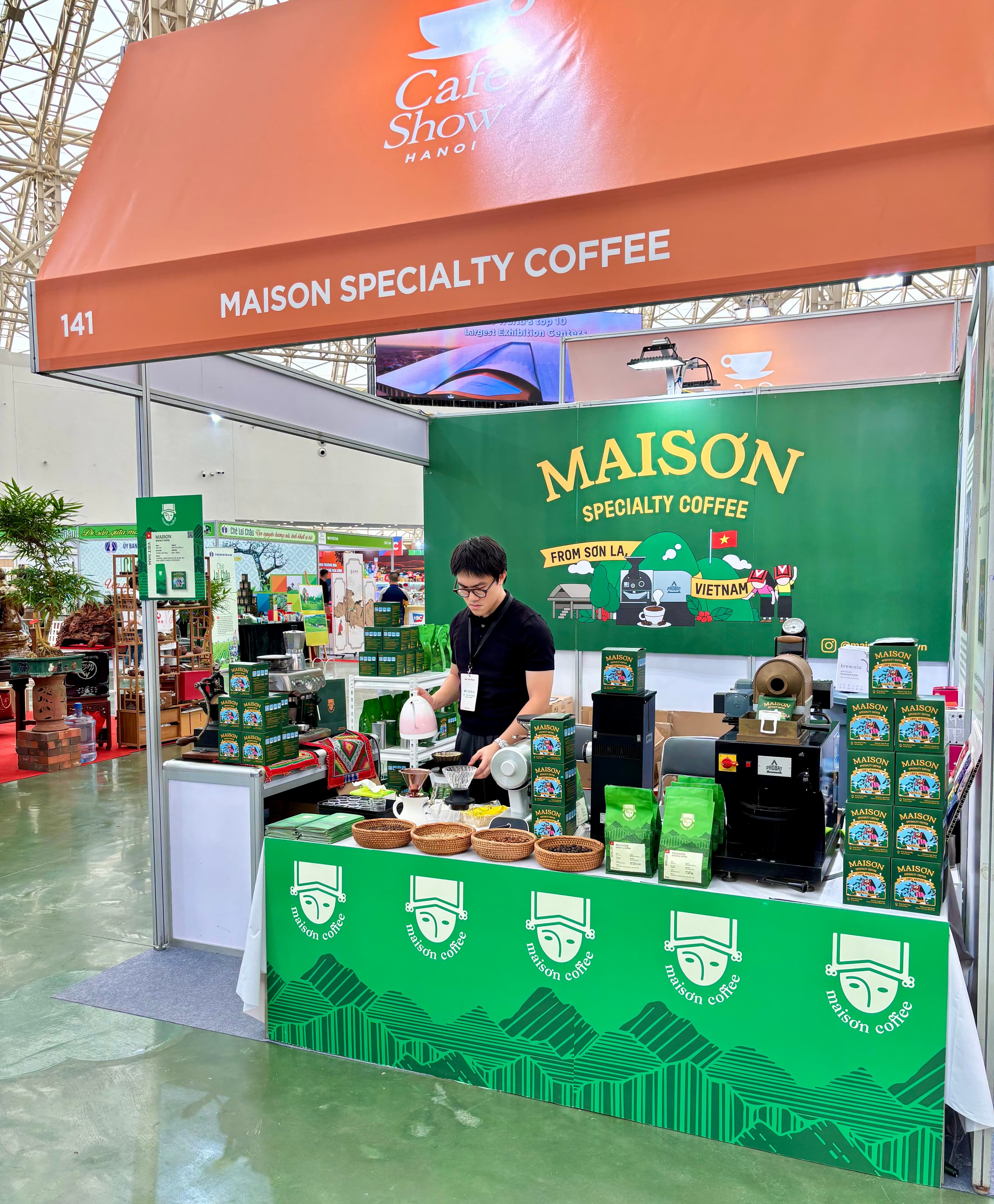 Maison Coffee's booth at Cafe Show Hanoi, with shelves of Maison Specialty coffee and a staff member preparing a pour-over